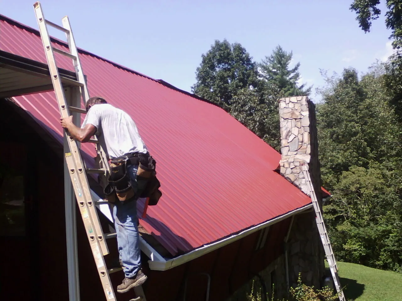 Roofer on a ladder beside a red metal roof and stone chimney — Soffit & Fascia Repair by Roofing Brothers in Dows